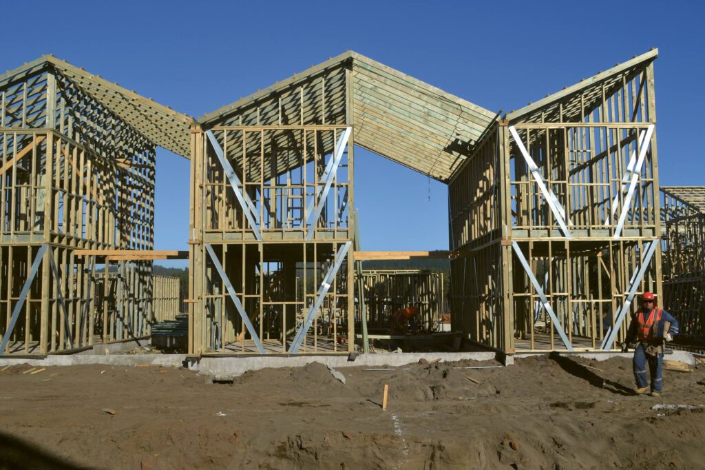 A wooden framework of homes under construction with sloped roofs, at a busy building site where workers in safety vests are active under a sunny blue sky.