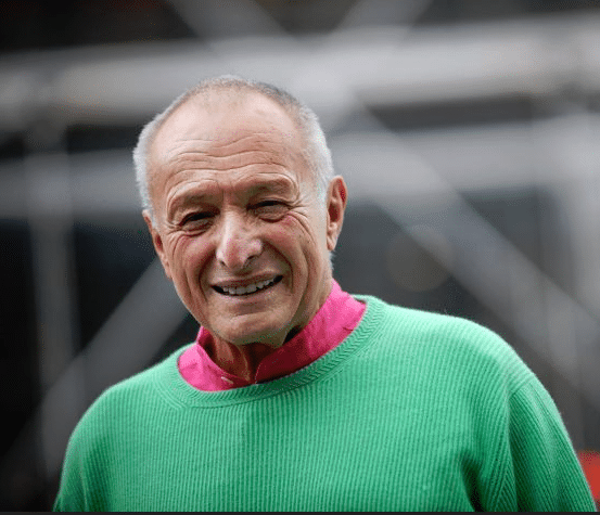 A man wearing a green sweater and a pink shirt, smiling broadly. Behind him is a modern architectural structure with glass surfaces and geometric lines