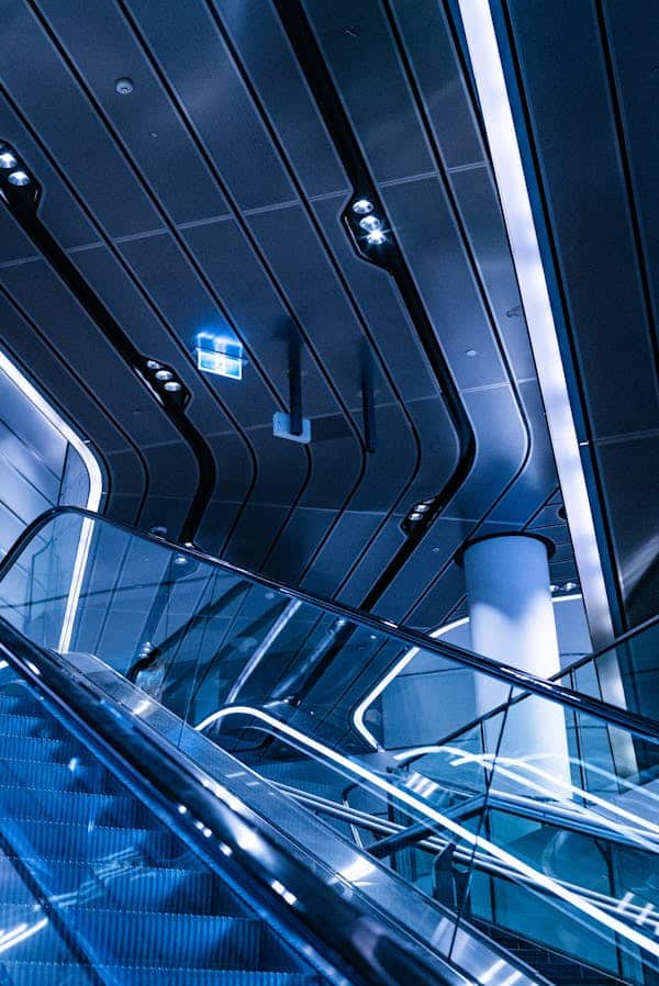 An interior view of a modern escalator within a building, showcasing a sophisticated design with materials like glass and metal. Soft blue lighting creates a modern, tech-inspired ambiance, complemented by illuminated guide lines on the walls