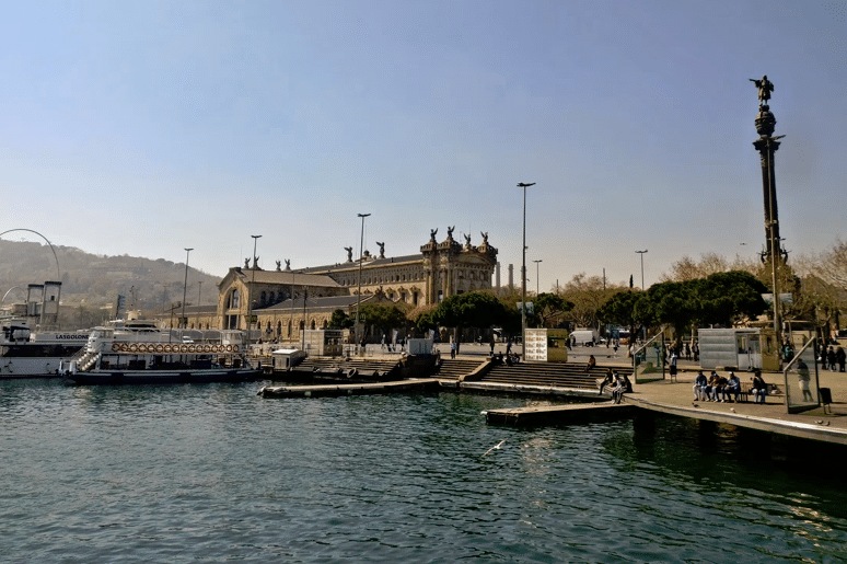 ميناء تاريخي في برشلونة، حيث تتوسطه نصب تذكارية ومباني قديمة تحمل طابعًا عريقًا، مع وجود سفن راسية على الماء وساحات للجلوس تستمتع بها الناس