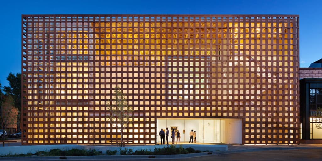 A modern building illuminated at night, featuring a grid of square windows on its reddish-brown facade. The structure has a lattice pattern that allows warm light to spill out, creating a glowing effect. In front of the building, a group of people stands near an open entrance, with small plants and trees surrounding the area.