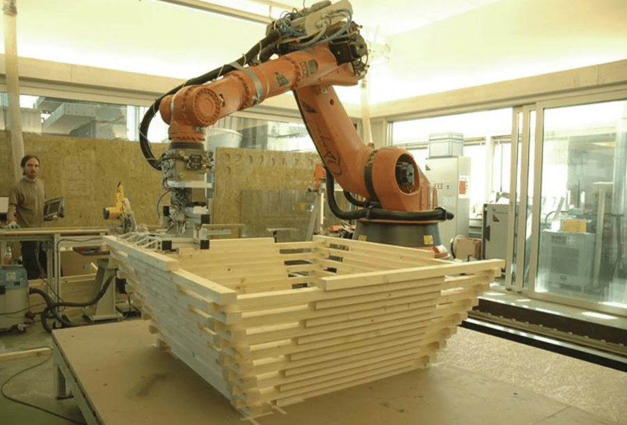 An industrial robot is being used to process wooden pieces in a manufacturing workshop, with a person in the background observing the operation