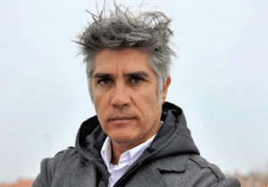 رجل يرتدي معطفًا أسود وقميصًا أبيض، ويبدو أنه في الهواء الطلق. شعره أشعث وألوانه مزيج من الرمادي والأسود،