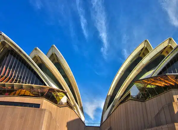 The iconic facade of the Sydney Opera House, featuring its distinctive and complex design with curved surfaces resembling seashells, set against a clear blue sky.