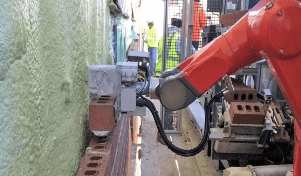 An orange industrial robot placing bricks on a wall, supervised by workers wearing safety gear.