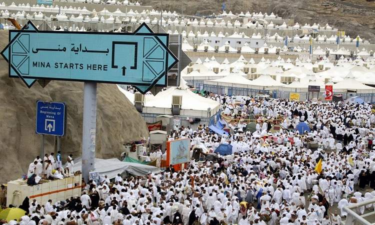 A large crowd of pilgrims dressed in the distinctive white attire for Hajj and Umrah walking along a busy path. In the foreground, there is a prominent blue sign that reads 'Mina Starts Here' in both Arabic and English, with additional directional signs indicating the beginning of Mina. The background shows thousands of white tents stretching across a vast area.