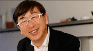 A man wearing white-framed glasses and a black suit, sitting at a table with his hands clasped together. He is smiling warmly, exuding confidence and approachability in a modern, professional environment
