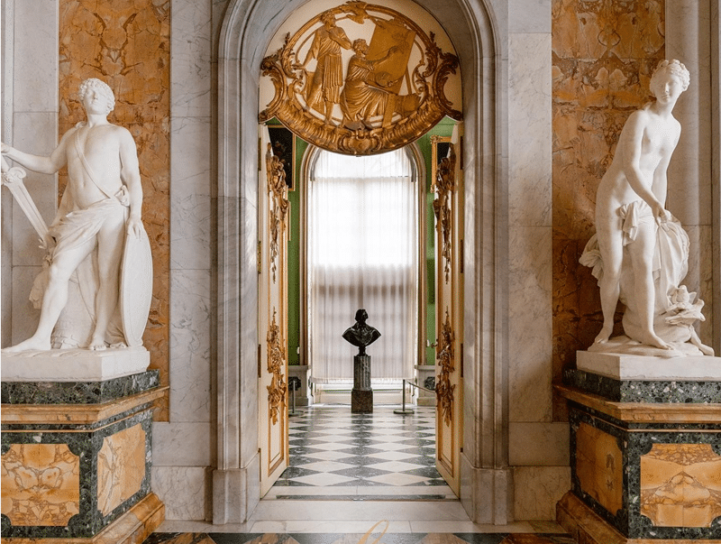 "An opulent interior hallway adorned with marble columns, statues, and intricate gold detailing, showcasing the luxurious use of marble in classical architecture."