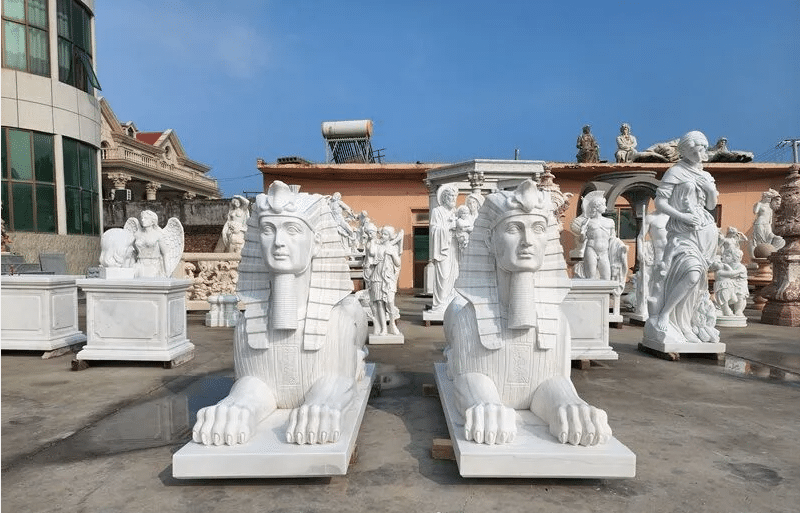 "A collection of marble sculptures, including sphinxes and classical figures, displayed outdoors in a sunny setting."