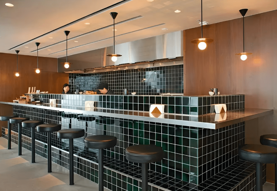 A modern restaurant featuring a long bar counter with leather stools underneath, surrounded by small black tiles that add a contemporary touch. Natural lighting enhances the space, complemented by simple hanging lights that provide a warm ambiance.