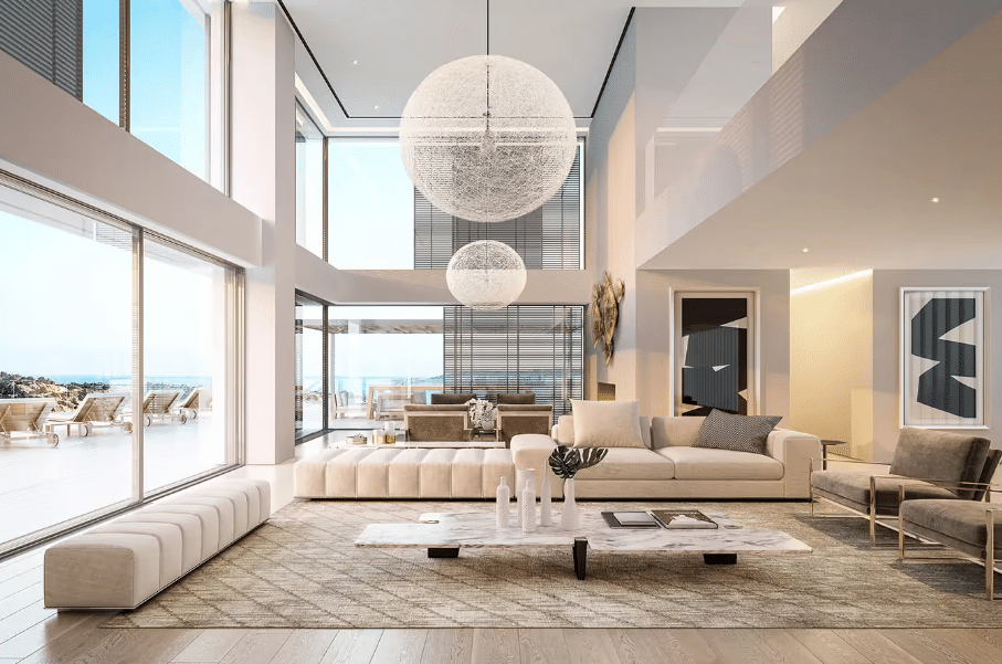 A modern living room with a sleek design, featuring a white sofa with dark cushions, a marble coffee table, and a wooden floor in soft tones. Tall walls with integrated lighting strips and large spherical pendant lights add an artistic touch. Floor-to-ceiling windows offer a serene view of the sea, enhancing the sense of openness and relaxation.