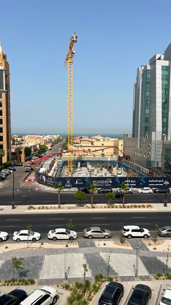 برج كرين ضخم يهيمن على المشهد في موقع البناء لبرج فهد في ألمحمرة، محيطًا بمجمعات سكنية وتجارية من عدة طوابق ونشاط حضري نشيط. الموقع محاط بشبكة زرقاء تحمل شعار البرج، مما يشير إلى مشروع هندسي رائد ضمن تطور الحدث الحضري السريع.