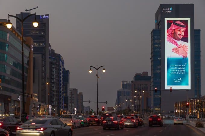 A nighttime view of Al-Khobar showcases a large digital billboard displaying a traditional figure in Saudi attire, surrounded by modern skyscrapers and illuminated streets. The red glow of traffic lights adds to the vibrant, active atmosphere of the city after dusk.