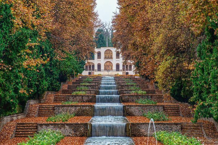 A tranquil garden featuring stone steps with a cascading waterfall, surrounded by trees with lush green leaves and golden autumn foliage.