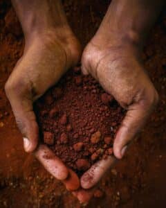 Human hands holding a handful of rich, reddish soil, the primary material used in rammed earth construction.