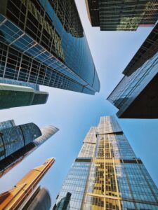 A stunning view of several towering skyscrapers captured from a low angle, making them appear to touch the clear blue sky. The buildings feature reflective glass facades that mirror the light and sky, creating a sense of height and modernity.