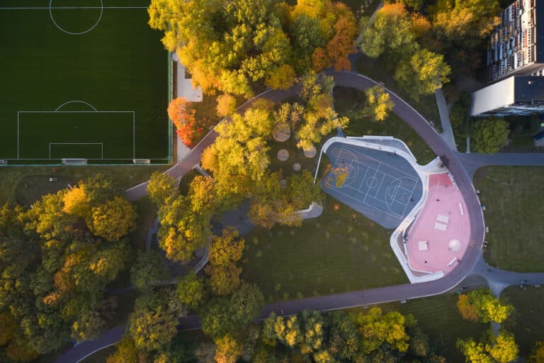 An aerial view of Oak Grove Park in Klaipėda, Lithuania, showcasing colorful sports courts surrounded by lush green and autumnal trees."