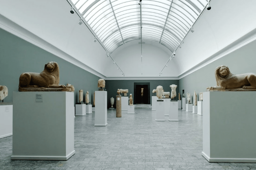 A museum gallery with a high, arched glass ceiling, featuring an array of ancient stone sculptures displayed on white pedestals in organized rows. The sculptures include animal and human figures, illuminated by natural light streaming through the glass roof.