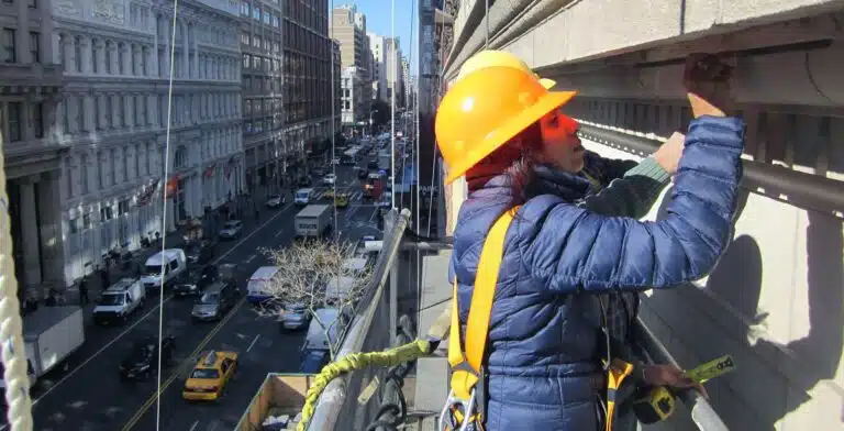 A technician wearing a safety helmet and harness conducting a facade inspection in New York City.