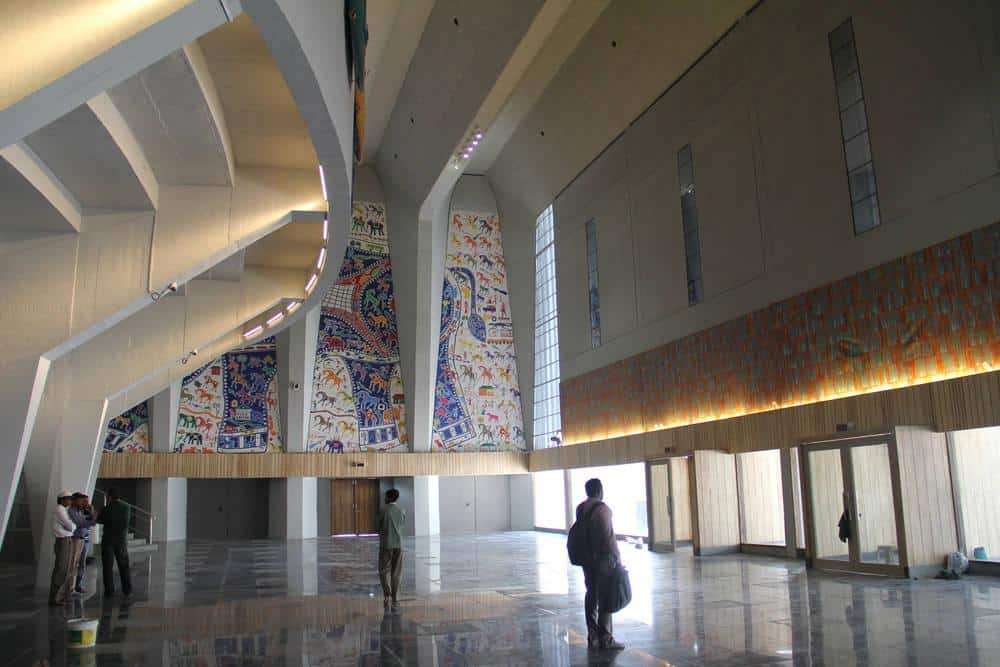 An indoor art exhibition in a modern building featuring large, colorful murals with vibrant designs, and people walking and enjoying the artwork.