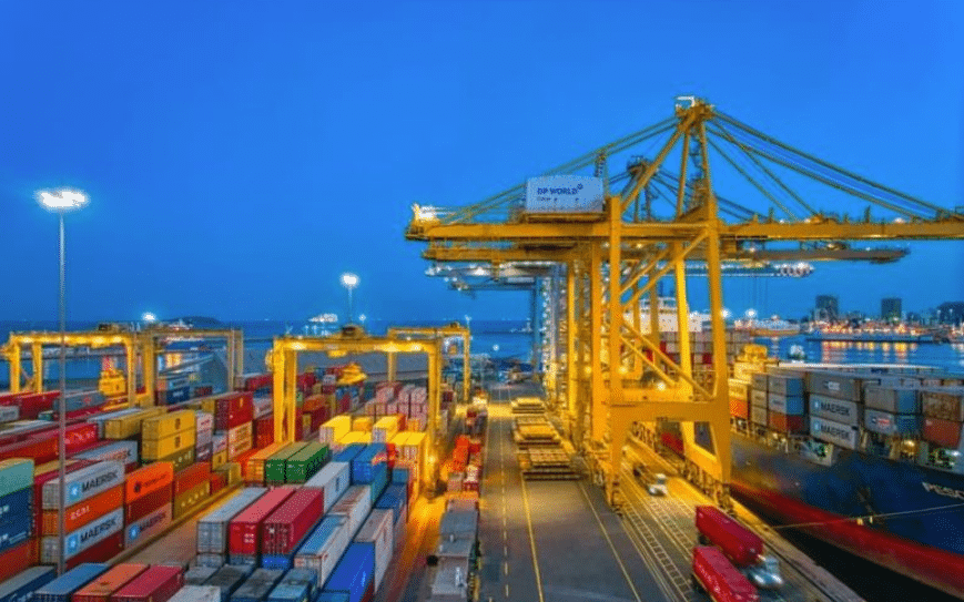 A bustling commercial port illuminated at night, with industrial cranes and colorful containers glowing under the lights, alongside a large cargo ship in the background