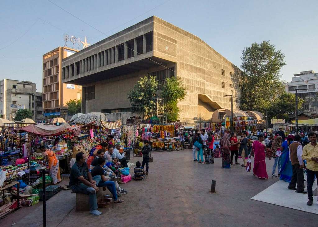 A bustling street market with people shopping and sitting in an open area in front of a modern building with flat roofs and concrete pillars.