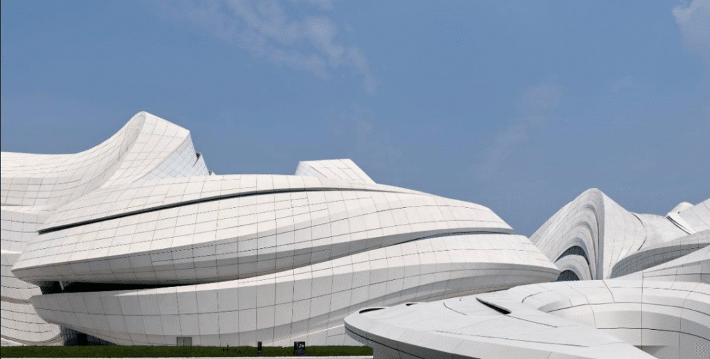 A modern architectural design of a building featuring curved and wavy surfaces covered with white panels, highlighting the unique shape of the structure. The blue sky in the background adds to the aesthetic appeal.