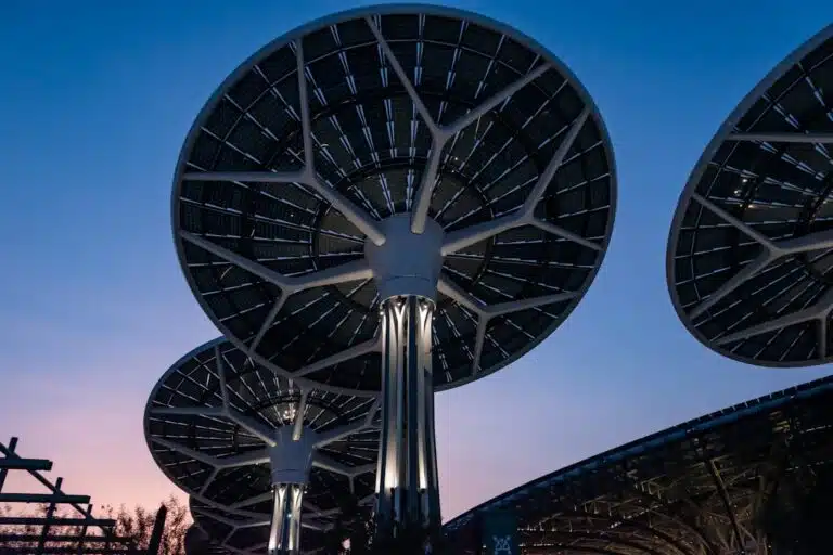 A tensile structure in the shape of a circular dome, composed of a network of cables and flexible membranes illuminated with soft internal lighting, set against a twilight sky with shades of blue and magenta.
