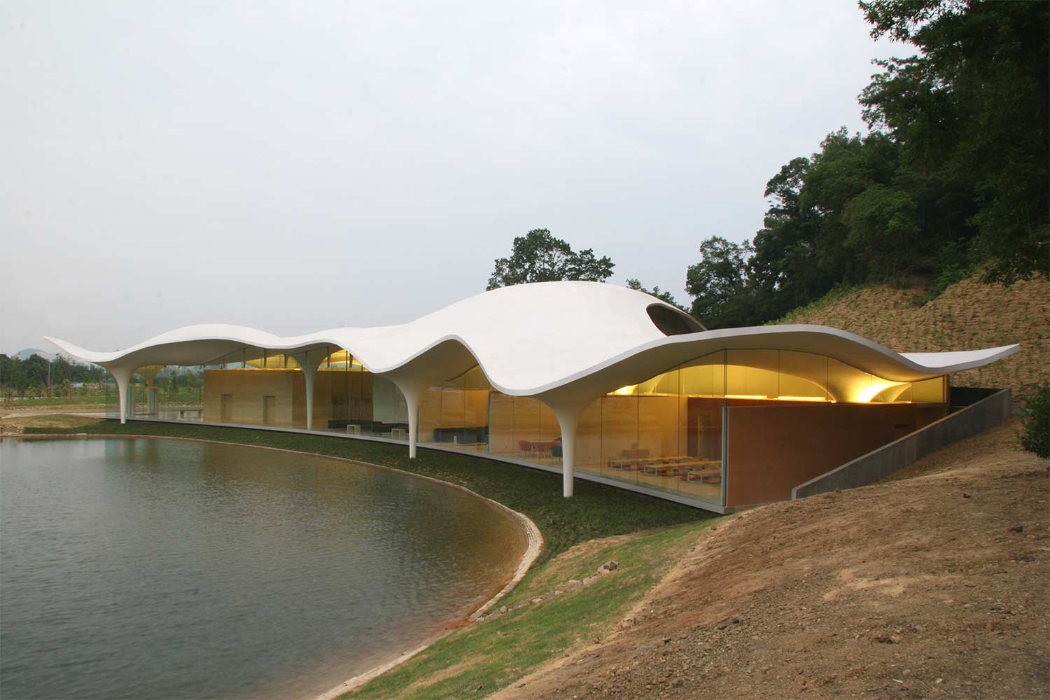 A sleek architectural design reflecting Toyo Ito's philosophy, featuring a smooth, flowing white structure along the edge of a lake, with warm interior lighting that blends nature and technology seamlessly."