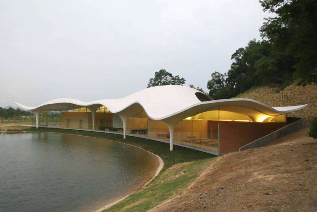 A sleek, modern building featuring a flowing, wave-like white roof that gracefully curves over the structure. The building is surrounded by a serene body of water and lush greenery, creating a harmonious blend between architecture and nature