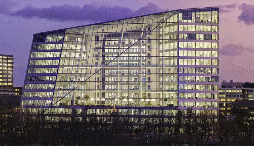 A modern glass building illuminated from within with warm lights during dusk, overlooking an urban cityscape.