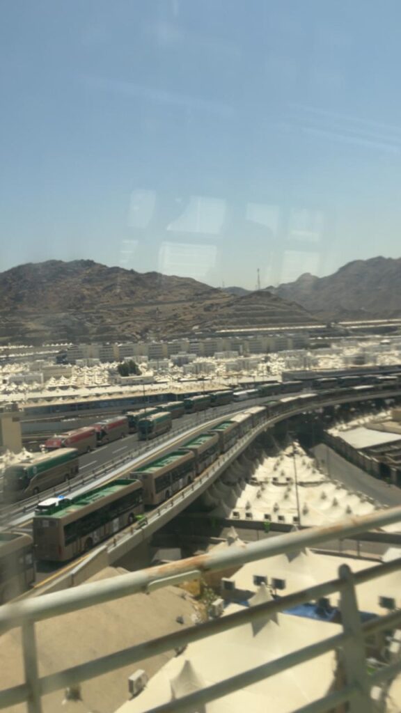 A top-down view showing buses moving on a bridge with white tents spread out below the mountains in the Hajj area.