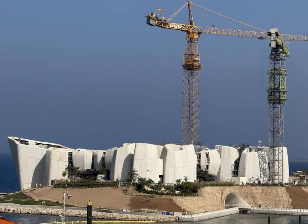 A striking modern building under construction, featuring a series of white, curved structures that blend seamlessly with the surrounding landscape. Tower cranes dominate the scene, indicating active development. The structure is situated near the water, with clear blue skies and distant mountains in the background, emphasizing the integration of architecture with nature.