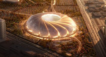 A futuristic stadium with a unique, cup-shaped design illuminated at night, surrounded by greenery and modern infrastructure.