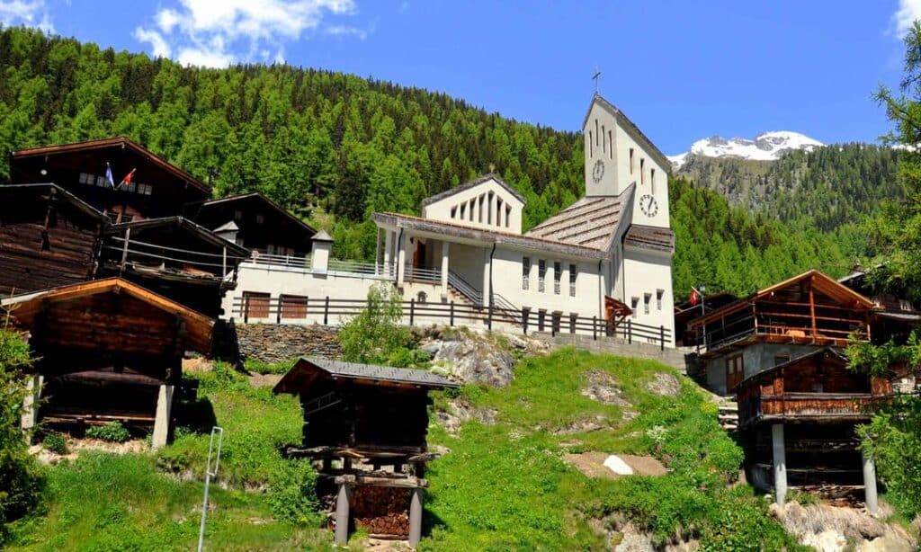 A white church stands amidst a small Swiss village, surrounded by traditional wooden houses and lush green mountains in the background.