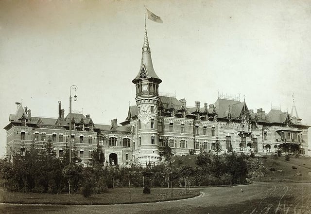 A majestic palace with intricate architecture, featuring a tall tower with a clock and a flag flying at the top, surrounded by lush greenery.