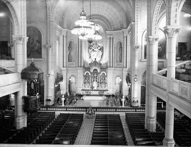 The interior of a grand church, featuring high arched ceilings adorned with intricate patterns and detailed moldings. Rows of wooden pews line the nave, leading to an ornate altar at the far end. A large chandelier hangs from the ceiling, casting light over the space, while religious statues and paintings decorate the walls and side altars.