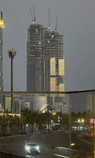 أبراج سمو في جدة تظهر تحت الإنشاء، حيث يلمع بعض الأضواء على الهياكل الحديدية والأسمنتية أثناء الليل المبكر. الواجهة البحرية مضاءة بأضواء الشوارع والمباني المجاورة، مع وجود ممرات للمشاة وأشجار على طول الطريق.