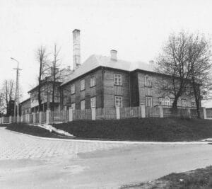 An old wooden nursing home with a simple facade, featuring evenly spaced windows and small chimneys, surrounded by a wooden fence and a small garden.