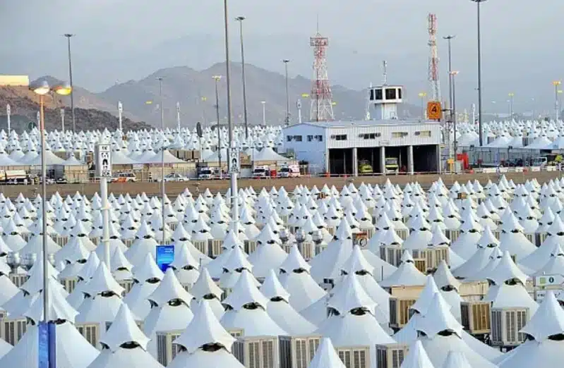 A wide view showing thousands of neatly arranged white tents in the Hajj area, with a building, towers, and distant mountains in the background.