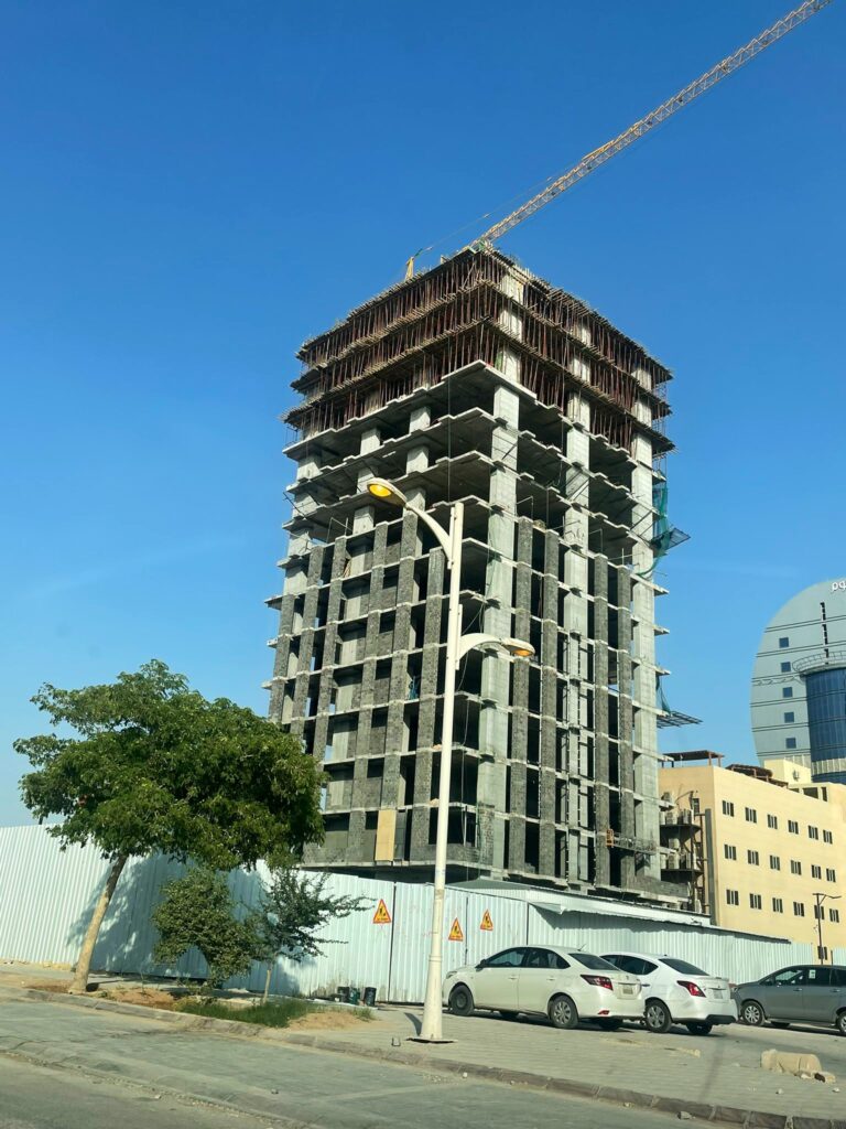 A tall, partially constructed concrete building under construction with scaffolding and a crane in the background. The structure is surrounded by urban elements like parked cars, trees, and nearby buildings.