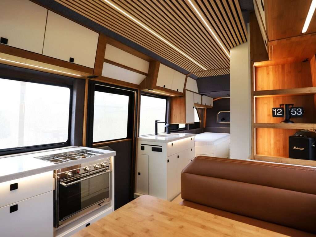 A modern kitchen in the Torsus Liberra bus, featuring integrated appliances like a stove and washing machine, with warm wooden lighting and built-in storage shelves, showcasing a functional yet luxurious design