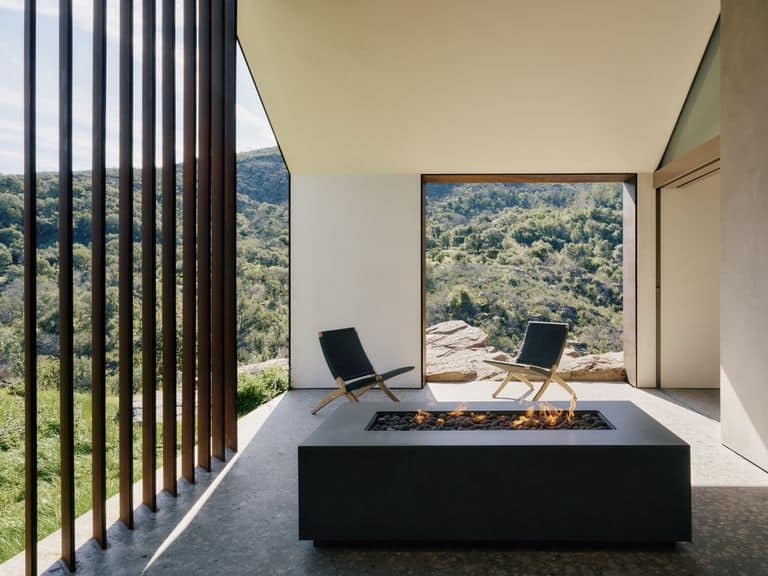 منظر داخلي يظهر منطقة جلوس مع كرسيين خشبيين أمام حوض نار صغير، ويطل على منظر طبيعي واسع للغابة عبر فتحات زجاجية كبيرة.