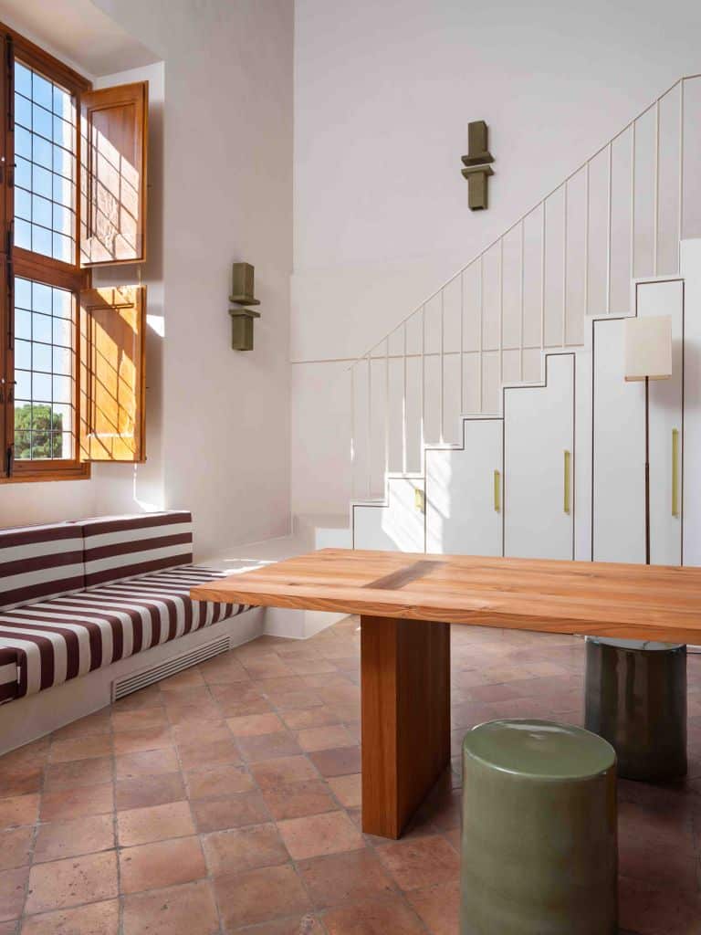 A bright room that blends traditional and modern elements, featuring a long striped bench against the wall, a large wooden table with simple design, and floor cushions made of wicker. The wooden-framed window adds warmth, while the white staircase with geometric lines brings a contemporary touch to the space.