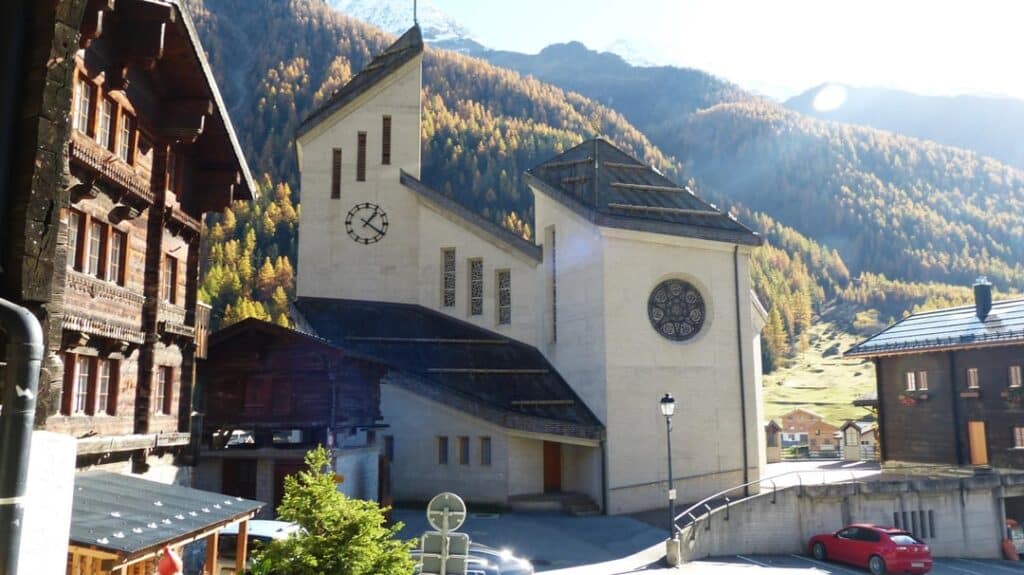 A simple, white church with a minimalist architectural design, situated in a Swiss village surrounded by traditional wooden houses and lush green mountains.