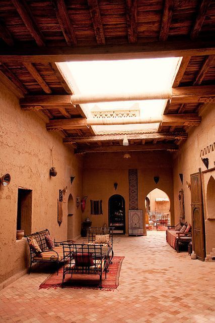 A traditional indoor courtyard featuring wooden furniture and earthy tile floors, with a wooden ceiling that includes a glass skylight allowing natural light to brighten the space, creating a serene and cozy atmosphere.