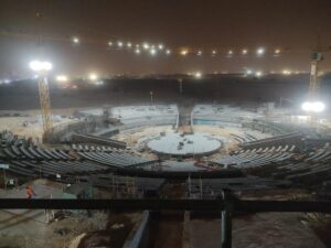 An image of an ongoing construction project at night, showcasing a massive architectural structure with a circular, tiered design. The site is illuminated by bright construction lights, and cranes are visible in the background, working on assembling various parts of the building.