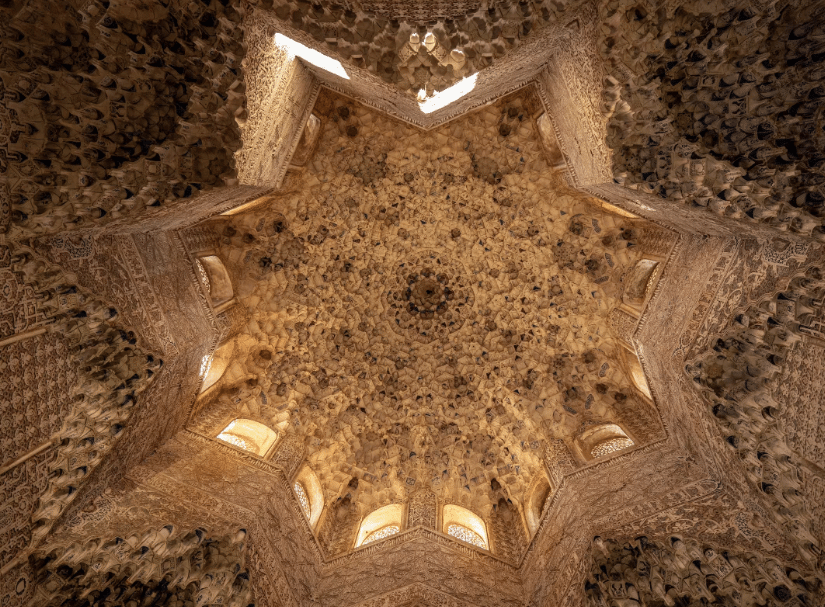 A beautifully decorated ceiling with intricate stone muqarnas, featuring geometric precision and glass windows allowing natural light to filter through.