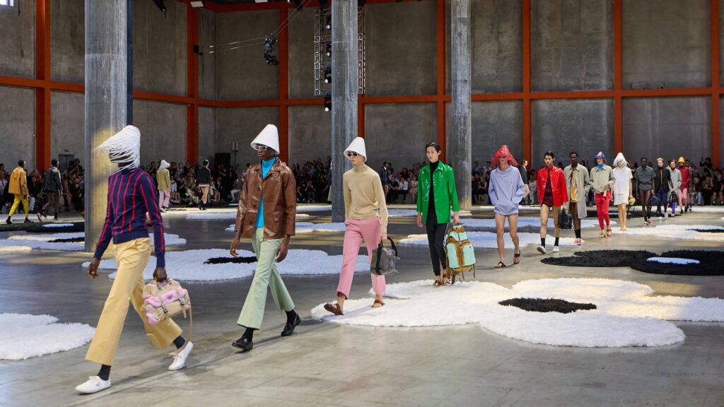 A group of models walking on an open runway, wearing colorful and unconventional fashion designs, with a large floral rug in the center adding visual interest to the industrial setting.