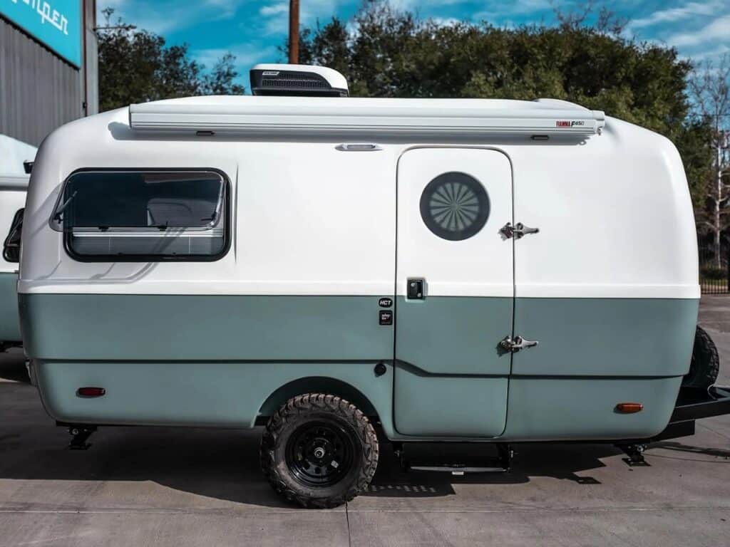 A compact camping trailer with a stylish design, featuring white and light blue colors and circular windows that add an aesthetic touch. The trailer is equipped with rugged tires suitable for off-road adventures.